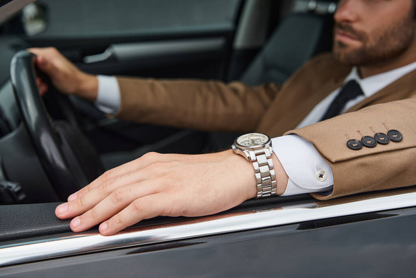 bijgesneden beeld van zelfverzekerde zakenman in beige pak en polshorloge rijden luxe auto op straat - Foto, afbeelding