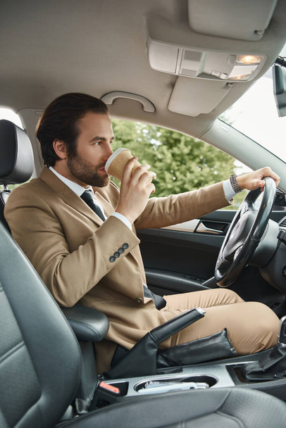 стильный бородатый бизнесмен в бежевом костюме пьет кофе, чтобы поехать и водить машину на городской улице - Фото, изображение