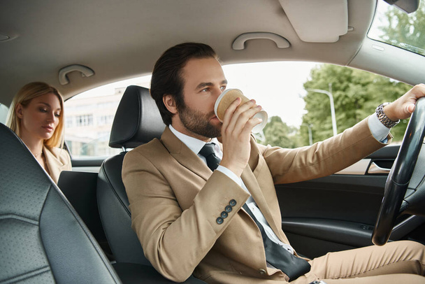 uomo elegante in abbigliamento formale bere caffè e guidare auto con donna d'affari bionda sul sedile posteriore - Foto, immagini