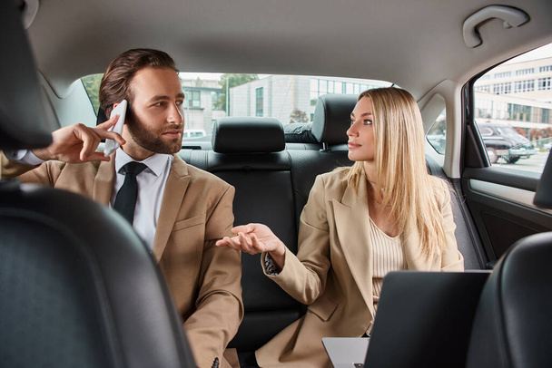 blonde businesswoman talking to handsome man in formal wear calling on smartphone in luxury car - Photo, Image