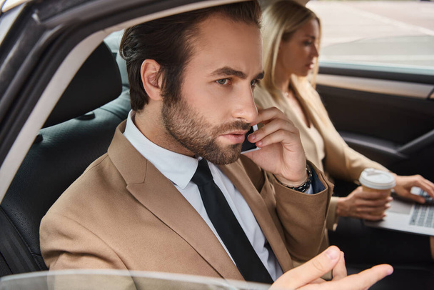 man in formal wear talking on smartphone near businesswoman with coffee to go and laptop in car - Photo, Image