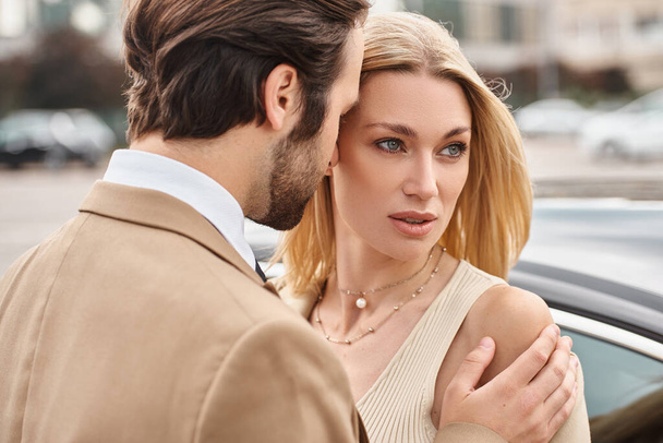 biznesmen obejmujący zmysłowy blondynka w formalnym stroju w pobliżu samochodu na ulicy miejskiej, przyciąganie - Zdjęcie, obraz