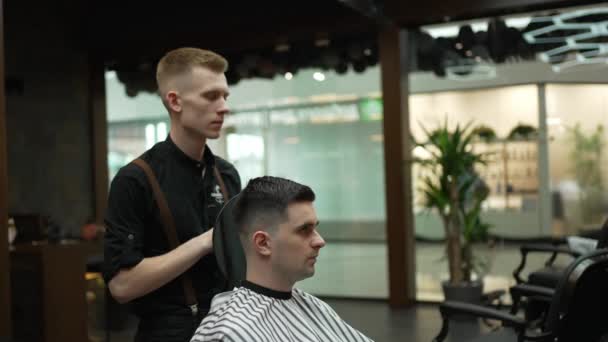 Перукар показує результат стрижки на спині голови для клієнта, який сидить у кріслі у дзеркалі - Кадри, відео