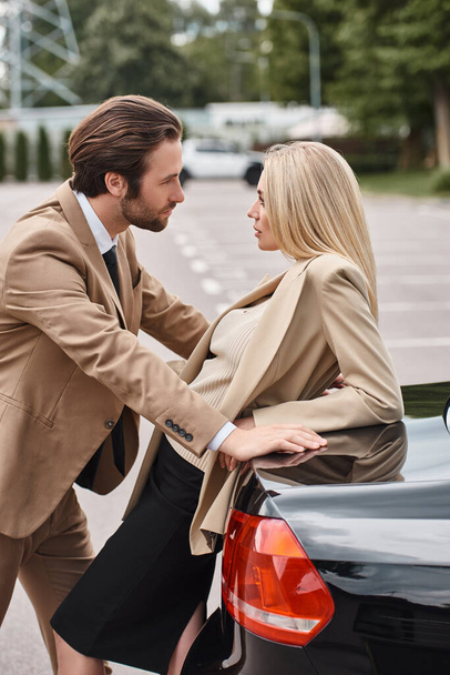 κομψός άντρας με επίσημη ένδυση κοιτάζοντας αισθησιακή ξανθιά επιχειρηματίας κοντά στο αυτοκίνητο στο δρόμο, έλξη - Φωτογραφία, εικόνα