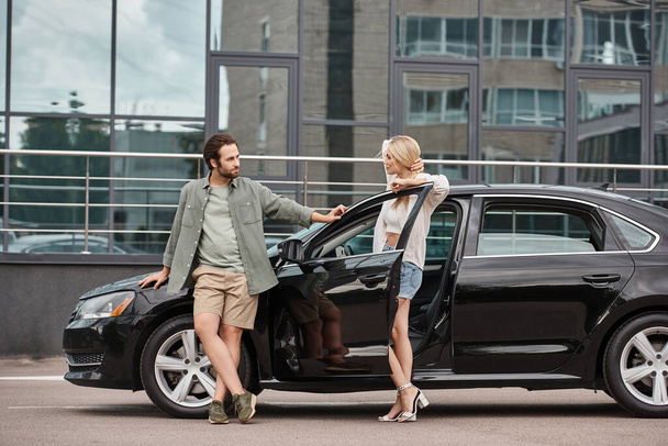 attractive couple in trendy casual clothes looking at each other near car on city street, affection - Photo, Image