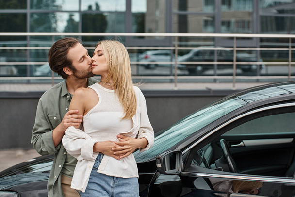 魅惑的なスタイリッシュなブロンドの女性を抱擁し,都市の通りで近代的な車の近くにキスしたひげ付き男 - 写真・画像