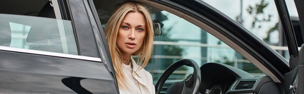 mujer rubia elegante y expresiva sentada en un coche moderno y mirando a la cámara, pancarta horizontal - Foto, Imagen