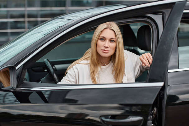 Blondine in trendiger Freizeitkleidung sitzt in modernem Auto und blickt auf der Straße in die Kamera - Foto, Bild