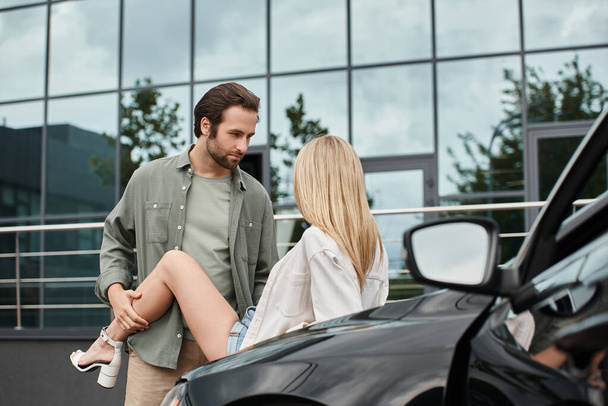 knappe bebaarde man in trendy casual kleding verleiden blonde vrouw in de buurt van auto op straat, genegenheid - Foto, afbeelding