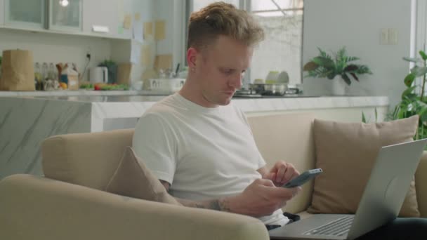 Közepes lövés a fiatal férfi ül kanapén otthon, a laptop és okostelefon használata közben online vásárlás vagy a számlák kifizetése - Felvétel, videó