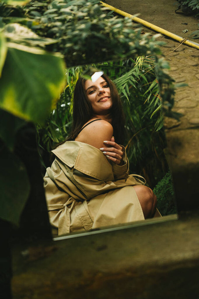 Mujer sexy en una gabardina beige posando en un jardín tropical. Concepto de tiro de moda - Foto, Imagen