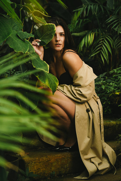 Sexy Frau im beigen Trenchcoat posiert in einem tropischen Garten. Konzept für Modeschießen - Foto, Bild