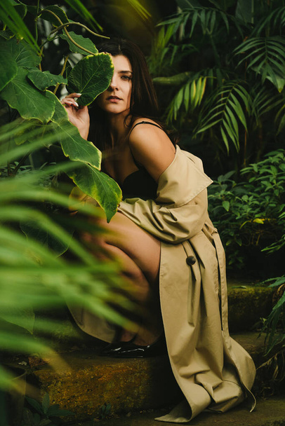 Sexy woman in a beige trench coat posing in a tropical garden. Fashion shooting concept - Photo, Image