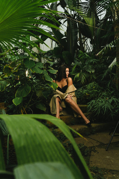 Tropik bir bahçede poz veren bej renkli trençkotlu seksi kadın. Moda çekim konsepti - Fotoğraf, Görsel