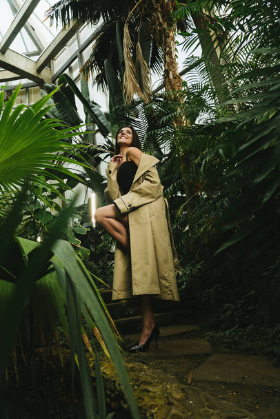 Sexy vrouw in een beige trench jas poseren in een tropische tuin. Modeschietconcept - Foto, afbeelding