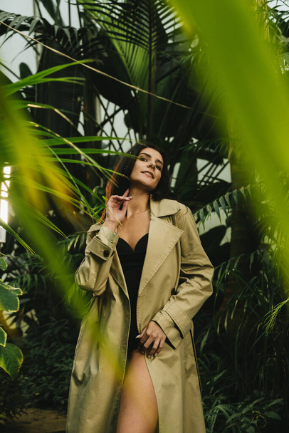 Сексуальная женщина в бежевом плаще позирует в тропическом саду. Концепция модной съемки - Фото, изображение