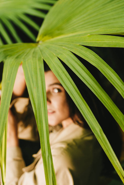 Сексуальна жінка в бежевому траншеї позує в тропічному саду. Концепція зйомки моди - Фото, зображення