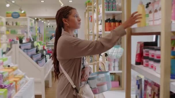 Colpo medio lungo di giovane donna afroamericana che mette lo shampoo nel carrello della spesa al negozio di cosmetici - Filmati, video