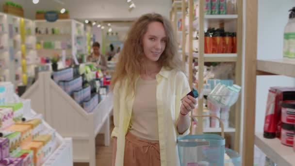 Portrait of young positive Caucasian female customer choosing moisturizer during shopping at cosmetics store, looking at camera - Footage, Video