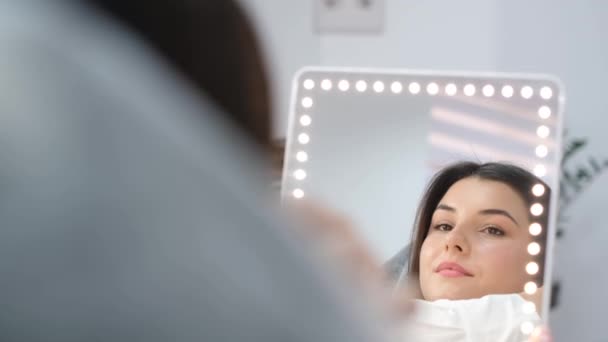 Красивая женщина смотрит в зеркало после косметических процедур. Уход за лицом у косметолога. - Кадры, видео