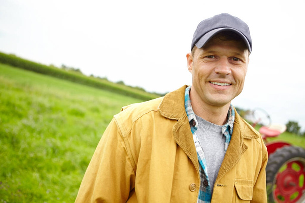 Rakastan työtäni. Muotokuva hymyilevä viljelijä pellolla traktorin pysäköity hänen takanaan - Copyspace - Valokuva, kuva
