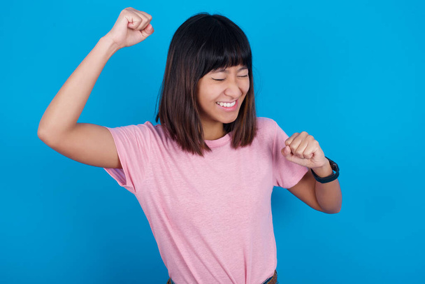 Atractiva joven asiática vistiendo camiseta sobre fondo azul celebrando una victoria golpeando el aire con sus puños y una sonrisa radiante. - Foto, Imagen