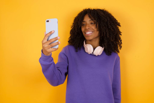 portrait of young expressive african american woman with headphones and smartphone on yellow background - Photo, Image