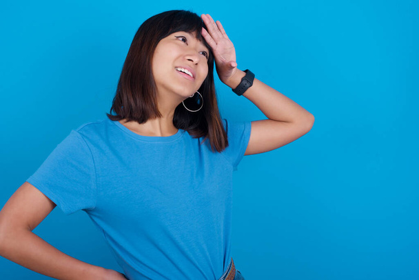 Asyalı genç bir kadın mavi arka planda t-shirt giyip alnına dokunuyor, şaşırtıcı bir şey duyuyor, iyi haberler aldığına seviniyor, rahatlıyor. Neredeyse başım belaya giriyordu.. - Fotoğraf, Görsel