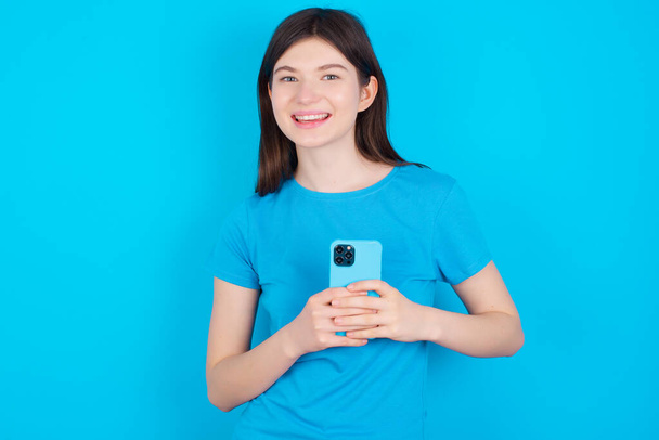 Uśmiechnięta młoda kaukaska dziewczyna w niebieskiej koszulce odizolowana od niebieskiego tła przyjazna i szczęśliwie trzymająca telefon komórkowy robiąca selfie w lustrze. - Zdjęcie, obraz