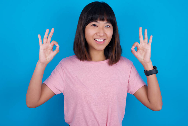 青い背景にTシャツを着用している若いアジアの女性は,両手でOKのサインを表現し,楽観的で陽気な表情を持っています. - 写真・画像