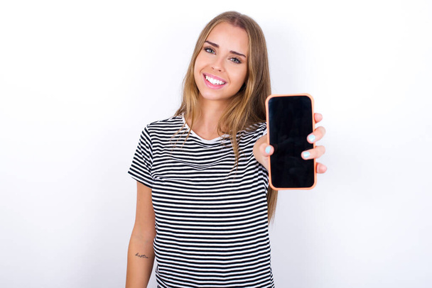 Очаровательная очаровательная красивая блондинка в полосатой футболке на белом фоне с современным устройством, показывающим черный экран смартфона - Фото, изображение
