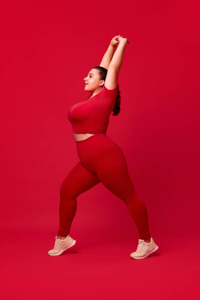 Beautiful overweight woman in comfortable sportswear training, stretching against red studio background. Concept of sport, body-positivity, weight loss, body and health care, self-acceptance - Photo, Image