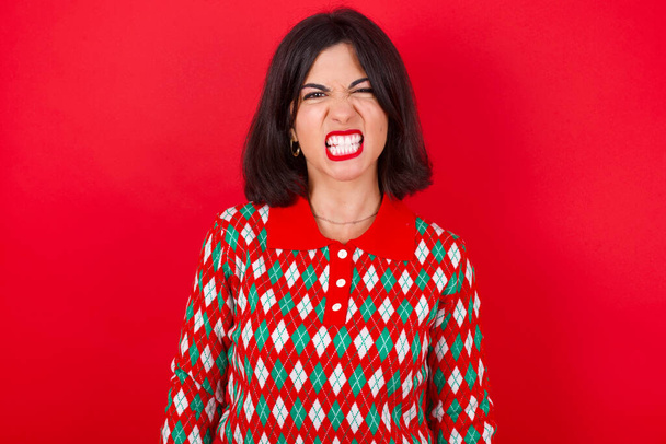 brunette Kaukasische vrouw dragen kerst trui over rode achtergrond houdt tanden gebald, fronst gezicht in ontevredenheid, geïrriteerd vanwege veel plichten. - Foto, afbeelding