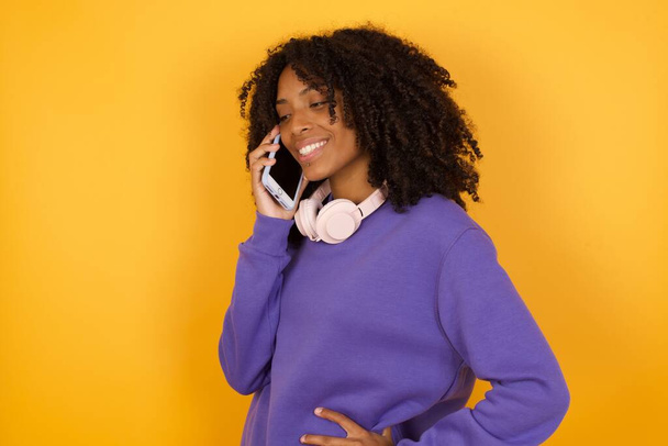 portrait de jeune femme afro-américaine expressive avec écouteurs parlant par téléphone sur fond jaune - Photo, image