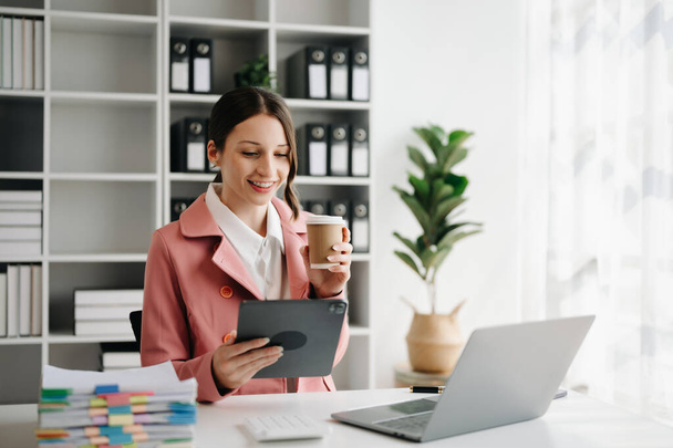 Empresária caucasiana bonita confiante usando tablet digital e computador portátil enquanto segura café no escritório moderno - Foto, Imagem