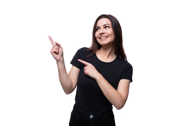 Jonge blanke vrouw met bruine ogen gekleed in zwarte kleren kijkt naar de zijkant en wijst met haar vinger. - Foto, afbeelding