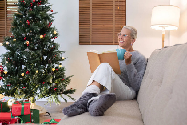Mulher aposentada A década de 60 passou as férias de Natal relaxando, desfrutando de uma xícara de chá e lendo um livro no sofá da sala de estar.. - Foto, Imagem