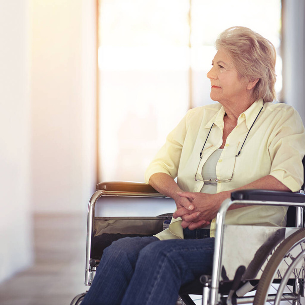 Κάνω ένα ταξίδι στη μνήμη. μια ηλικιωμένη γυναίκα που δείχνει σκεπτική ενώ κάθεται σε μια αναπηρική καρέκλα στο σπίτι - Φωτογραφία, εικόνα