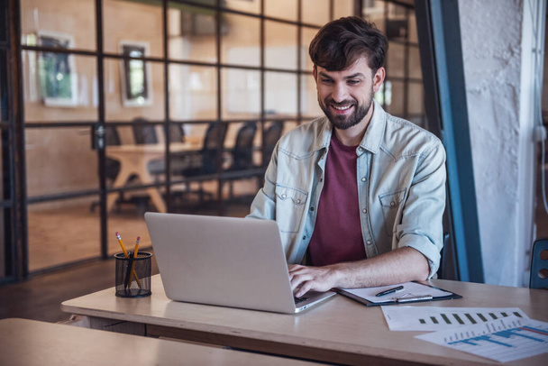 Bonito empresário barbudo em roupas casuais está usando um laptop e sorrindo enquanto trabalhava no escritório
 - Foto, Imagem