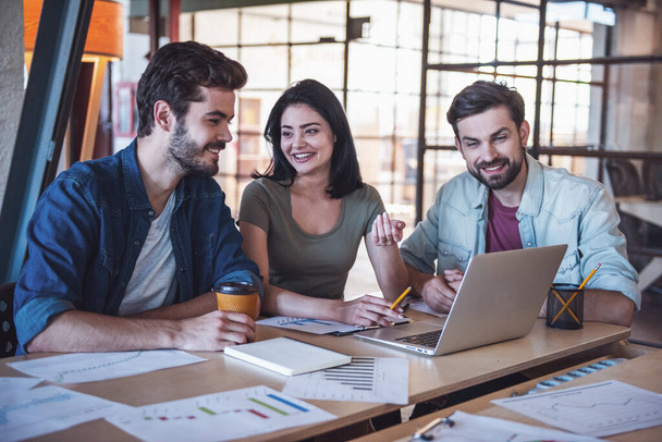 Belos jovens empresários em roupas casuais estão usando um laptop, conversando e sorrindo enquanto trabalham no escritório
 - Foto, Imagem