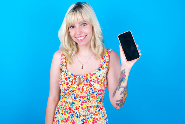 Mosolygó kaukázusi lány virágos ruhát visel elszigetelt kék háttér mutatja üres telefon képernyőn. Reklám és kommunikációs koncepció. - Fotó, kép