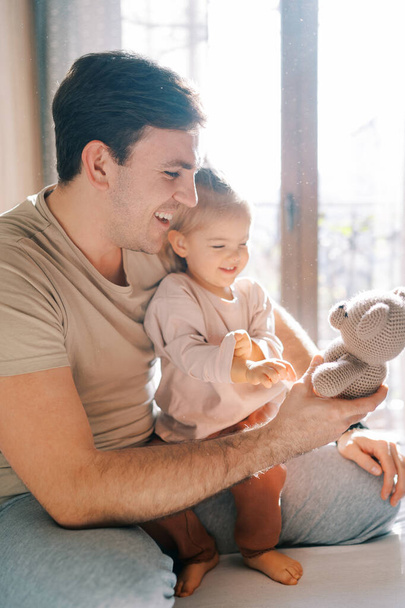 Little smiling girl sits on the lap of a laughing dad with a teddy bear in his hand. High quality photo - Fotoğraf, Görsel