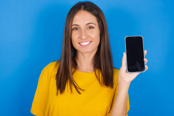 Uśmiechnięta piękna młoda kobieta pokazuje pusty ekran telefonu. Koncepcja reklamy i komunikacji. - Zdjęcie, obraz