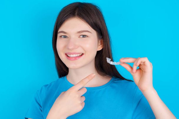 Mavi tişört giyen beyaz bir genç kız mavi stüdyo arka planında izole edilmiş ve elinde görünmez bir hizalama aleti var. Diş sağlığı ve güven kavramı. - Fotoğraf, Görsel