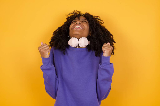 retrato de joven mujer afroamericana expresiva con auriculares sobre fondo amarillo celebrando - Foto, imagen