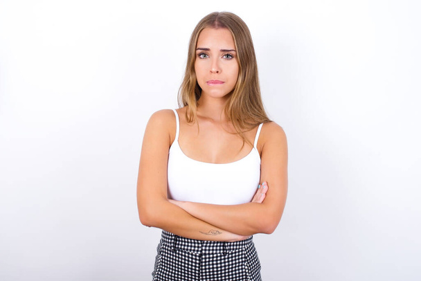 Young Caucasian girl wearing white tank top on white background biting his mouth and looking worried and scared crossing arms, worry and doubt. - Photo, Image