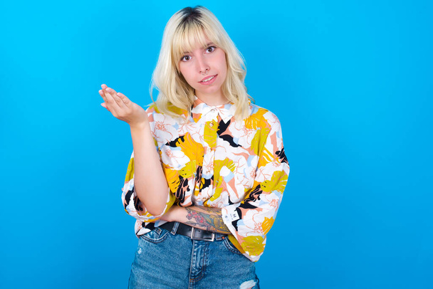 Studio shot of frustrated Caucasian girl wearing floral shirt isolated over blue background gesturing with raised palm, frowning, being displeased and confused with dumb question. - Φωτογραφία, εικόνα