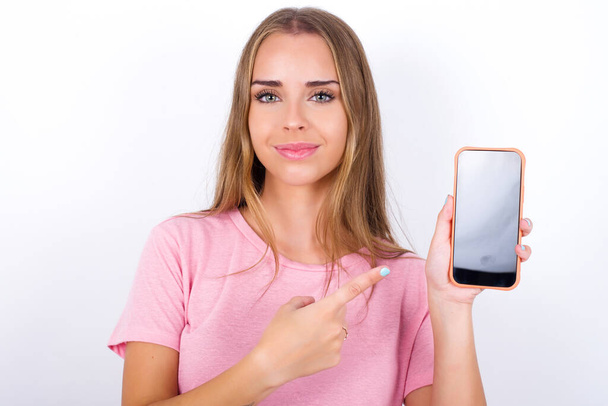 Lächelndes junges kaukasisches Mädchen in rosa T-Shirt auf weißem Hintergrund, das auf den leeren Telefonbildschirm zeigt. Werbe- und Kommunikationskonzept. - Foto, Bild