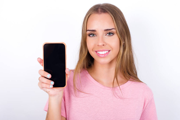 Lächelndes junges kaukasisches Mädchen in rosa T-Shirt auf weißem Hintergrund, das den leeren Telefonbildschirm zeigt. Werbe- und Kommunikationskonzept. - Foto, Bild