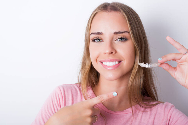 Young Caucasian girl wearing pink T-shirt on white background holding an invisible aligner and pointing at it. Dental healthcare and confidence concept. - Foto, afbeelding